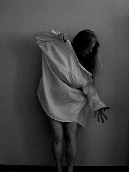 A woman is standing in front of an indoor wall trying to dress up in a bright shirt backwards. One arm is holding the shirt up in front of her and she is looking down on her hand that has just passed through the other sleeve. You can see part of her face in profile, the rest is hidden by her long hair hanging down. Her legs are bare.