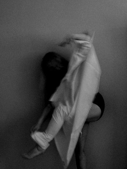 A woman is trying to put a shirt on but instead of putting her arms in the sleeves she is trying to get it on both backwards and with one arm in one sleeve and her left leg in the other one. Her left arm is holding the shirt up and she is leaning forwards since she can´t stand straight with the shirt put on like that. Her hair is haning down, hiding her face and her but is partly visible. She wears panties but her standing leg is bare. This image is also a bit blurry because of her motion during exposure.