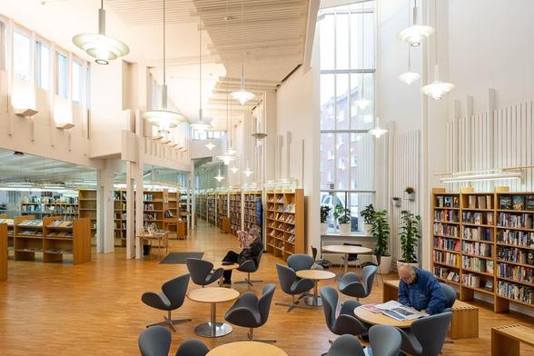 Interior of a modern library with white walls and blonde wood furniture. There is cafe seating in the foreground, with people reading newspapers.