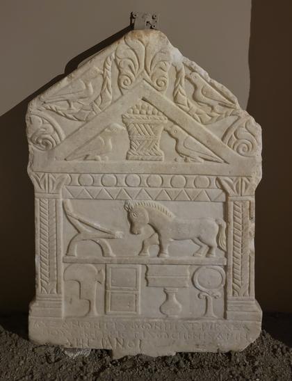 Marble tombstone, on which is carved a cross-section of a classical temple (columns and triangular roof). Inside the roof are carved two doves pecking at a basket of food. On the second level of the "house" is a horse and an unidentified furniture-like object. On the first level are other objects resembling containers, mirrors, books, etc. At bottom, below the house, is the Greek inscription.