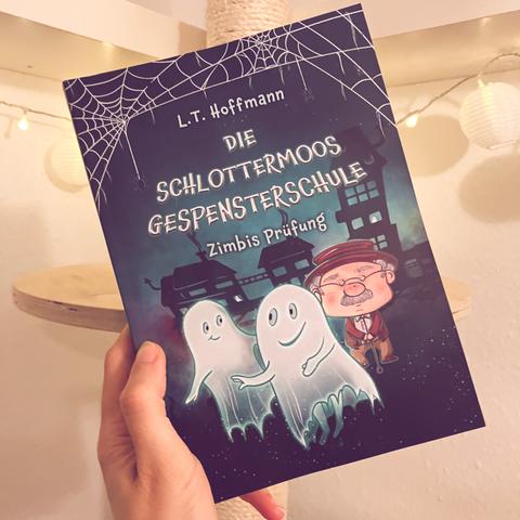 Eine Hand hält das Kinderbuch "Die Schlottermoos Gespensterschule" von L. T. Hoffmann in die Höhe.