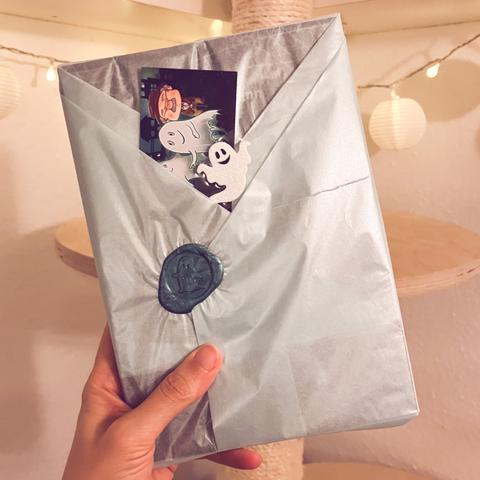 Eine Hand hält ein noch verpacktes Buch in die Höhe. Aus dem gefalteten Einpackpapier schaut ein Lesezeichen und ein kleiner Geist-Sticker heraus.