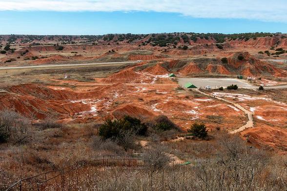 A landscape featuring rolling red hills stretches toward the horizon beneath a partly cloudy sky. Sparse vegetation and a dirt path wind through the arid terrain, with a few structures visible in the distance. Photography by Debra Martz