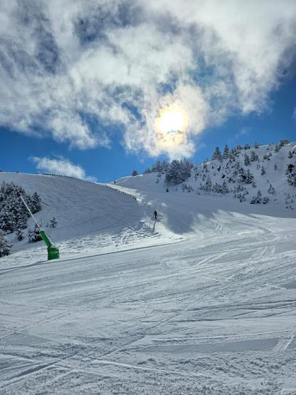 Picture of a ski slope with just 1 skier. The sun is shining and the sky is blue with some white clouds. The slope is not very steep and there are some snowy mountains in the background.