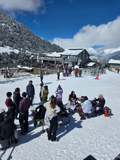 A group of people in ski- and snowboard gear resting around tables at the side of a ski slope. The people look happy and the sun is shining. The sky is blue with some big white clouds. Some partially snow covered mountains are visible in the background.
