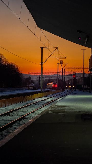 A train traveling along snowy tracks at sunset, with a warm orange sky and silhouettes of overhead electric lines and signals.