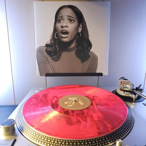 A transparent pink vinyl record sits on a turntable. Behind the turntable, a vinyl album outer sleeve is displayed. The front cover shows Kilo Kish looking off camera in surprise.