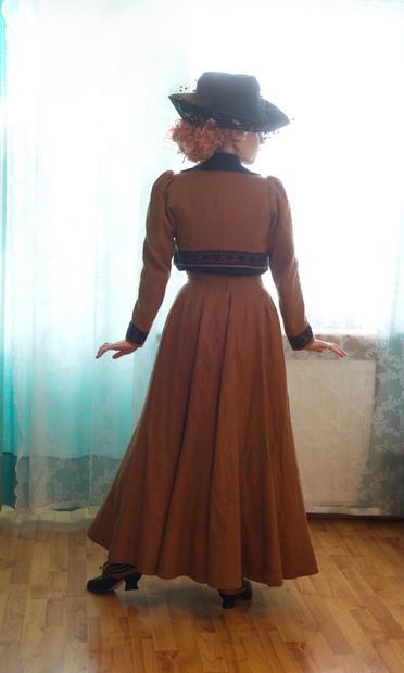 Back view. The skirt falls in beautiful smooth ripples, being nearly a circle skirt at the back, and the high waist of it sits just below the hem of the jacket, a little peek of both snatched waist and black blouse peeking out. It's a rather dynamic look. The jacket back sits nicely, but is of slightly the wrong width, thus messing with how the sleeves fall... While there is plenty of range of motion, there's a bunch of bunching up at the back of sleeve, and it would look much better in motion and with arms bent at the elbow, instead of this rather like a musical number jazz hands at the side pose. Hat looks cute. Red curly hair in a messy do spills out from under it.