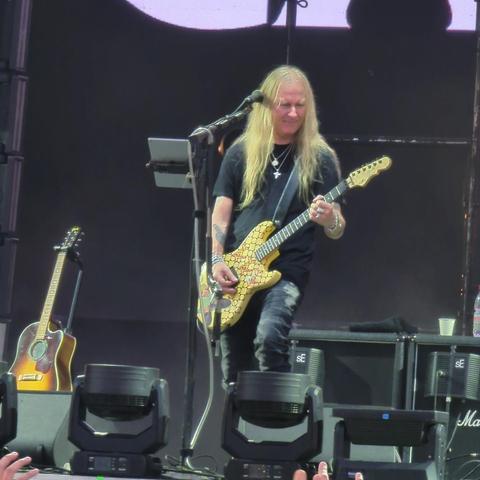 Jerry Cantrell luce cabello largo y rubio mientras está de pie en un escenario, tocando la guitarra eléctrica amarilla con un diseño personalizado. Lleva una camiseta negra y pantalones oscuros. En el fondo, se pueden ver amplificadores de sonido, focos y una guitarra acústica. Foto de su presentación en Fauna Primavera 2024 en Santiago de Chile.