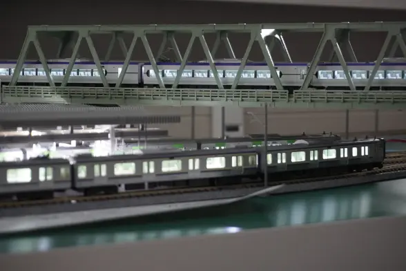 Model train set-up with a train station and a train bridge going over it. There is a train currently driving on the bridge. The station has multiple platforms and also has a train in the front.