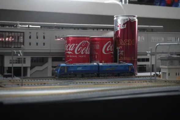 A model train set-up up with several tracks and a cargo train in front, as well as a big elevated station in the back. There are some drinking cans in-between the tracks in the front and the station in the back as model of silo's.