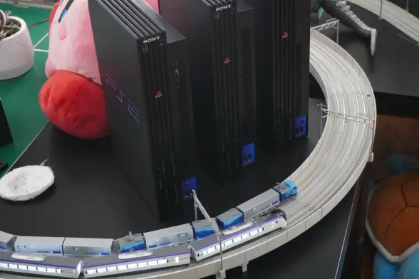 A big bend of the model train layout with 2 trains driving over the bend right now. The bend is right at "console city" a small skyline of 3 PS2's. Also in the background is a Kirby plushy.
