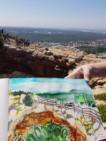 A relatively loose but colorful sketch of a land formation which looks like a natural bridge. The sketch is being held up by an arm in front of the actual landscape. Some of the natural bridge formation is visible there, with people standing on it, marveling at the view.