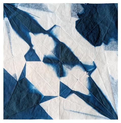 A piece of white paper which shows creases from being folded into an origami form - in this case a 'crane / orizuru' - and exposed as a cyanotype. This means it has blue geometric patterns all over it - what part of the paper was the head of the crane is top left, wings top right, bottom left, and tail. In the centre is a blurred blue x shape