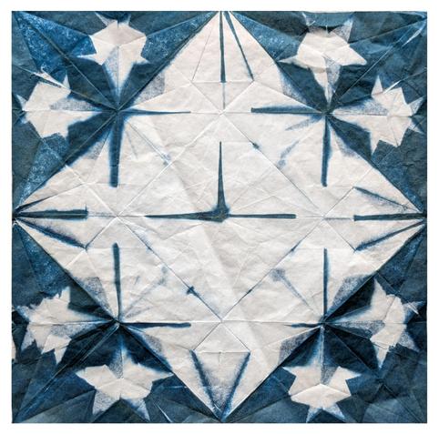 A piece of white paper which shows creases from being folded into an origami form - in this case a partially completed complex model of a sun - and exposed as a cyanotype. This means it has blue geometric patterns all over it - a central white diamond extends to the centre of all sides, the four large corners are mostly blue in odd triangles with white areas shaped like random stars