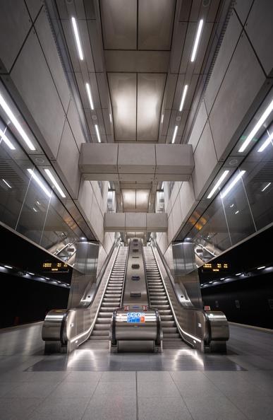 A symmetrical set of escalators in the tube station "Battersea Power Station". The architecture is sleek and industrial, featuring geometric concrete panels and bright overhead lighting. The reflective surfaces and metal railings contribute to a futuristic and minimalist aesthetic. Electronic signboards displaying train schedules are visible in the background. The station appears empty, emphasizing its clean and structured design.