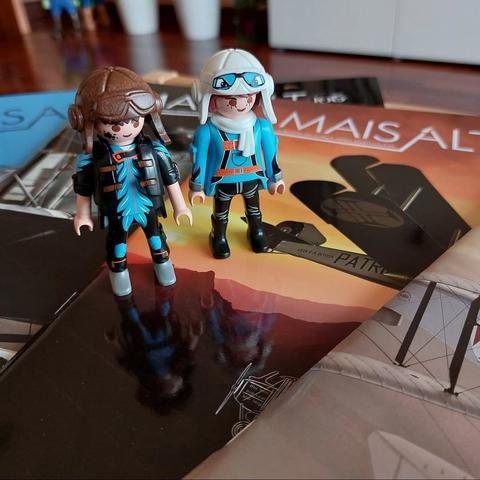 Dois pilotos antigos (capacete com óculos) da playmobil, homem e mulher, ambos de azul com capacete castanho e branco respectivamente, em cima de algumas revistas sobre aviação