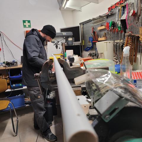Das Bild zeigt eine Werkstatt mit einer gut ausgestatteten Werkzeugwand im Hintergrund. Lars DL1STO mit dunkler Jacke, schwarzer Mütze und Brille arbeitet konzentriert an einem langen, weißen Rohr, das in einem Schraubstock eingespannt ist. Er scheint eine handwerkliche Tätigkeit durchzuführen, möglicherweise Bohren oder Bearbeiten des Rohrs.

Die Umgebung ist gut organisiert, mit zahlreichen Werkzeugen wie Schraubendrehern, Zangen und Bohrern an der Wand. Zudem sind verschiedene Arbeitsmaterialien, Kunststoffbehälter und eine Bohrmaschine auf der Werkbank zu sehen. Ein Erste-Hilfe-Schild hängt an der Wand, was auf eine sicherheitsbewusste Arbeitsumgebung hinweist.

Insgesamt vermittelt das Bild den Eindruck einer präzisen handwerklichen Arbeit in einer gut ausgestatteten Werkstatt.