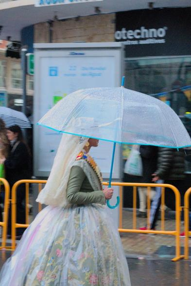 Una mujer vestida con el traje de fallera, vestido tradicional de valenciana, en la ofrenda a la Virgen, con paso ligero debajo de un paraguas que le cubre medio rostro ya que está lloviendo fuerte.