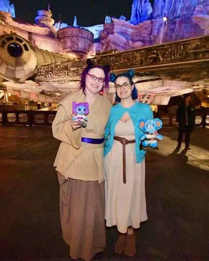 This image features two women posing together at Disneyland's Star Wars: Galaxy’s Edge, dressed as characters from the Star Wars franchise.

The woman on the left has dark hair styled in a bun and wears glasses, a tan robe, and brown pants. She holds a small blue creature with large ears and a long tail in her right hand. The woman on the right has short black hair and also wears glasses, along with a white dress and a blue cape draped over her shoulders. Her left arm is wrapped around the other woman's shoulder.

In the background, a large spaceship dominates the scene, while the dark sky above suggests it was taken at night.
