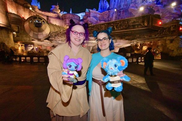 The image depicts two women standing side-by-side, each holding a stuffed animal. The woman on the left has purple hair and is dressed in a tan robe, while the woman on the right has blue hair and wears a white dress with a brown belt.

In her left hand, the woman on the left holds a small, pink cat plushie adorned with a bow around its neck. Meanwhile, the woman on the right cradles a light blue teddy bear in her left arm, which appears to be wearing an orange shirt and has a distinctive orange ear tag.

The background of the image features large metal object resembling a spaceship visible behind the women. The overall atmosphere suggests that the photo was taken at a themed event or convention, possibly related to science fiction or fantasy.