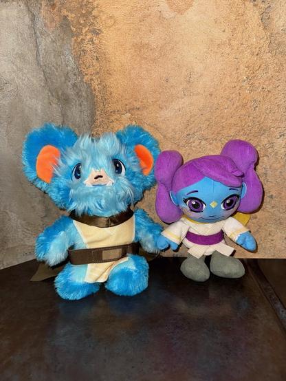 The image features two plush toys situated against a wall.

On the left is a blue bear-like creature with large ears, sporting an orange ear tip and a white belly. It wears a brown belt around its waist and has a small pink nose. The creature's fur appears to be made of soft, fuzzy material.

To the right stands a purple-haired doll with a white dress featuring a purple stripe across her chest. Her hair is styled in two large buns on top of her head, adorned with yellow accents. A gold necklace encircles her neck, and she wears gray shoes.

The background consists of a tan wall with darker brown spots, likely due to the texture of the wall or shadows cast by the toys. The overall atmosphere suggests that this image is intended for use as an advertisement for these plush toys.