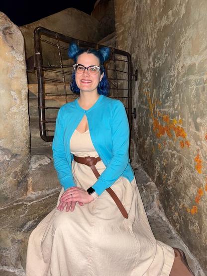 The image features a woman seated against a stone wall, her head turned slightly to the left as she gazes directly at the camera.

She has short blue hair styled in two buns on either side of her head. Her attire consists of a beige dress paired with a bright blue cardigan and a brown belt around her waist. The woman's hands are clasped together in front of her, resting on her lap.

The stone wall behind her is adorned with vibrant orange graffiti, while a black metal railing is visible to the left. The overall atmosphere suggests that this photograph was taken at night or indoors, as evidenced by the dark background and subtle lighting.
