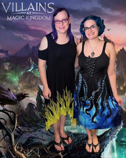 The image features two women standing together, posing for a photograph. The woman on the left has dark hair styled in a ponytail and is dressed in a black dress adorned with yellow flames at the hem. Her counterpart on the right boasts vibrant blue hair and wears a black tank top paired with a blue skirt featuring an octopus design.

In the background, a fantastical landscape unfolds, complete with towering mountains, lush greenery, and a majestic castle situated atop one of the peaks. The sky above is painted with hues of pink and purple, adding to the dreamlike quality of the scene.

At the top left corner of the image, the text "VILLAINS AT MAGIC KINGDOM" is prominently displayed in white letters.