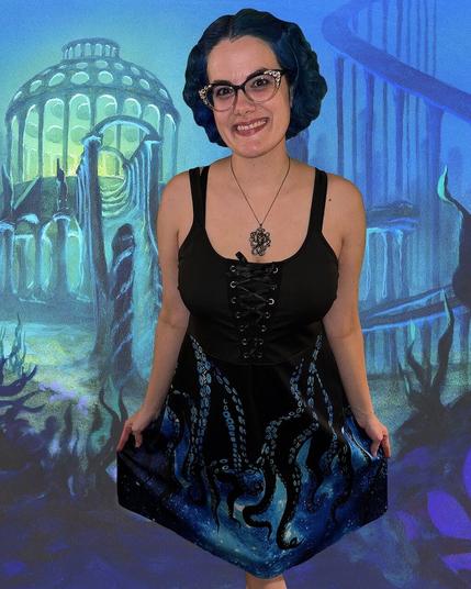 The image features a woman standing against a vibrant blue background, adorned with an underwater scene. She is dressed in a black tank top with lace-up details at the front, paired with a knee-length skirt that showcases an intricate design resembling tentacles from the deep sea. Her dark hair is styled in loose waves and falls just below her shoulders.

In the foreground, the woman's attire takes center stage, with the lace-up details on her tank top adding a touch of elegance to her overall appearance. The skirt's unique design draws attention to its intricate pattern, which appears to be inspired by oceanic creatures.

The background of the image is dominated by a bright blue hue, evoking an underwater atmosphere that complements the woman's attire perfectly. The presence of seaweed and other aquatic elements adds depth and context to the scene, creating a sense of immersion for the viewer.

Overall, the image presents a captivating visual narrative, with the woman's outfit serving