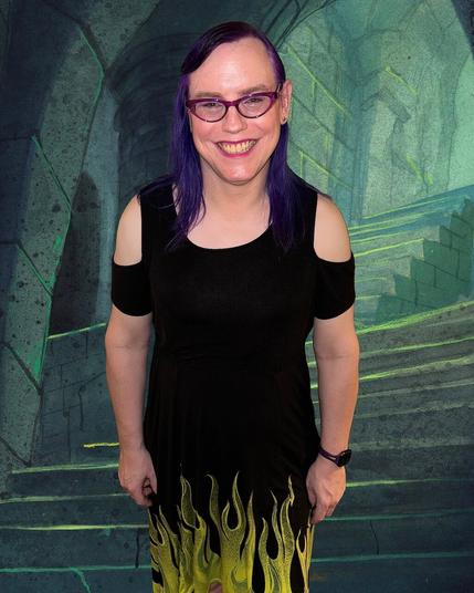 The image features a woman standing in front of a stone wall, likely an archway or doorway. She has long purple hair and is dressed in a black cold-shoulder top adorned with yellow flames at the hem. The background consists of large grey stones arranged in a herringbone pattern, with some green moss growing on them.