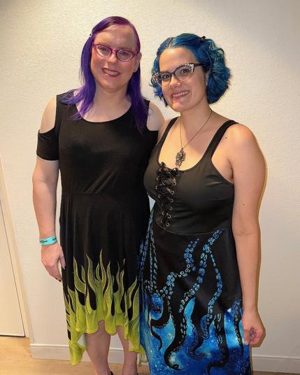 The image features two women standing side-by-side against a white wall background, posing for the camera.

Both individuals have distinctive hairstyles: one has purple hair, while the other sports blue locks. They are dressed in black dresses adorned with vibrant flame designs at their hemlines, adding a pop of color to the overall scene. The woman on the left wears a dress with yellow flames, whereas her counterpart on the right dons a dress featuring blue flames.

The women's attire is complemented by matching accessories: both wear glasses and necklaces, while the woman on the left also sports a teal bracelet on her wrist. Their facial expressions are neutral, conveying a sense of calmness as they pose for the camera.
