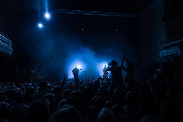 Black adn blue letterbox format photo of a music concert. Foggy background in blue adn the 2 guitarists in sillouette