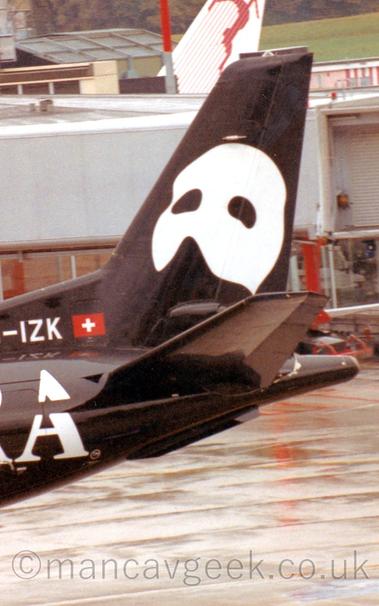 Close-up of the black rear fuselage and tail of an airliner taxiing from right to left.
The partial registration "-IZK" is visible on the base of the tail, above a large white letter "A" on the rear fuselage.
There is a large, white partial mask, designed to cover the upper face, on the tail.
Damp concrete apron fills the bottom of the frame in the background, with a grey terminal building behind the tail.