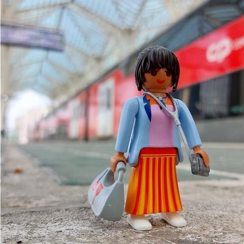 Mulher playmobil com cabelo castanho pele morena, casaco azul, saia listas vermelha e amarela torrada, com mala de mão azul, sorrindo,  na plataforma do comboio vermelho