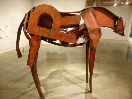 In an art gallery, a figure of a horse made from chunks of red-painted scrap steel.