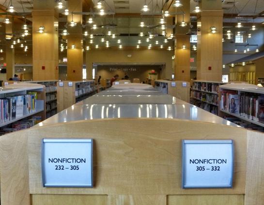 In a library, the endcap of a shelf unit for "Nonfiction", with many lights in the ceiling above, some reflecting from the top of the shelf.