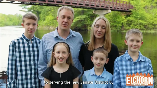 A clean cut very white family, well dressed, standing in front of a river with a hovering bridge and lots of greenery on its banks. There are four children spanning many ages, all minors.