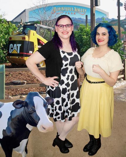 The image features two women standing side-by-side, posing for the camera. The woman on the left has purple hair and is dressed in a black cardigan over a cow-print dress, paired with white tights and black shoes. Her counterpart on the right boasts bright blue hair and wears a cream-colored blouse tucked into a yellow skirt, complemented by a black belt and black boots.

In the foreground, a small plastic cow statue is visible to the left of the women. Behind them, a large mural adorns the wall, depicting a serene landscape with trees, mountains, and a bright blue sky. A yellow construction vehicle is parked in front of the mural, adding a pop of color to the scene.

The overall atmosphere of the image suggests that it was taken at an amusement park or theme park, where the women are likely enjoying themselves on a sunny day.