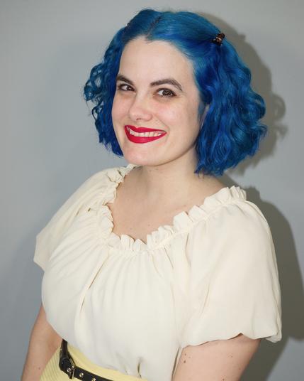 The image features a woman with striking blue hair styled in loose waves, complemented by a white blouse with ruffled edges around the neckline. Her attire consists of a yellow skirt paired with a black belt featuring silver studs. The background is a plain gray wall, providing a neutral setting for the subject.