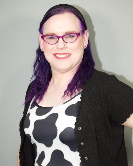 The image features a woman with long dark purple hair, styled straight and parted down the middle, wearing red-framed glasses and a black cardigan over a white shirt adorned with large black cow spots.

In the foreground, the woman is prominently displayed against a plain off-white wall. Her attire consists of a black cardigan worn over a white shirt featuring large black cow spots, complemented by red-framed glasses and long dark purple hair styled straight and parted down the middle. The background is a plain off-white wall, providing a neutral backdrop for the subject.

Overall, the image presents a clear and detailed portrait of the woman, with her distinctive hairstyle and attire standing out against the simple yet effective background.