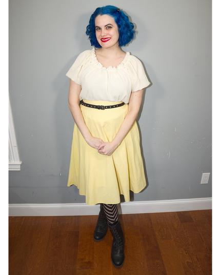 The image features a young woman standing against a gray wall, showcasing her vibrant blue hair styled in loose waves that cascade down to her shoulders. She is dressed in a white blouse with short sleeves and a V-neckline, paired with a yellow skirt that falls just above the knee. A black belt cinches at her waist, adding a touch of sophistication to her outfit.

The woman's attire is completed by black tights featuring horizontal stripes, which add a playful pop of contrast against the solid colors of her top and skirt. Her feet are clad in black boots with low heels, providing a subtle elevation without compromising comfort or style.

In the background, the gray wall provides a clean and neutral backdrop for the subject, allowing her colorful attire to take center stage. The overall atmosphere of the image is one of casual elegance, as if the woman has just stepped out of a vintage-inspired photoshoot.