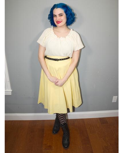 The image features a young woman standing against a gray wall, showcasing her vibrant blue hair styled in loose waves that cascade down to her shoulders. She is dressed in a white blouse with short sleeves and a V-neckline, paired with a yellow skirt that falls just above the knee. A black belt cinches at her waist, adding a touch of sophistication to her outfit.

The woman's attire is completed by black tights featuring horizontal stripes, which add a playful pop of contrast against the solid colors of her top and skirt. Her feet are clad in black boots with low heels, providing a subtle elevation without compromising comfort or style.

In the background, the gray wall provides a clean and neutral backdrop for the subject, allowing her colorful attire to take center stage. The overall atmosphere of the image is one of casual elegance, as if the woman has just stepped out of a vintage-inspired photoshoot.