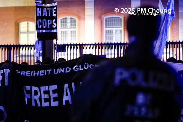 Vor der Justizvollzugsanstalt Moabit fand eine Zwischenkundgebung statt. Hier auf dem Bild ist ein Polizist zu sehen, ebenso ein kleines Stangentransparent mit der Aufschrift "Hate Cops".