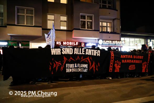 Die Demonstration bei der Auftaktkundgebung am U-Bahnhof Turmstraße. Hier sind zwei schwarze Banner zu sehen, auf einem steht "Wir sind alle Antifa. Feuer & Flamme dem Bullenstaat" und auf dem anderen "Antifa bleibt Handarbeit".