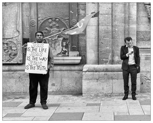 Two men in front of an old church with ornate doors. On the left, a man is holding a sign that states, "JESUS IS THE LIFE JESUS IS THE WAY JESUS IS THE TRUTH." He appears earnest and is looking directly at the camera. On the right, there is another man, dressed in a suit, who is engrossed in reading something on his smartphone, seemingly unaware or indifferent to the presence of the man with the sign. The scene captures a juxtaposition of religious proclamation and modern disengagement, set against a backdrop of historical architecture.