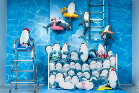 A playful swimming pool-themed IKEA display featuring numerous BLÅHAJ shark plushies arranged in a creative scene. Against a blue water-effect backdrop, the sharks are organized to create the illusion of a pool party. Several sharks wear colorful flower garlands, while others float with pool toys - one relaxes in a yellow duck inner tube and another with a pink flamingo float. One shark sits on a lifeguard chair on the left side, wearing a red cap. A metal pool ladder is mounted on the right side of the display with sharks climbing up it. The bottom portion shows multiple sharks stacked together like stadium seating or pool steps, all facing forward to create a unified aquatic tableau. The entire display cleverly transforms the iconic blue sharks into swimmers enjoying a summer pool day.​​​​​​​​​​​​​​​​