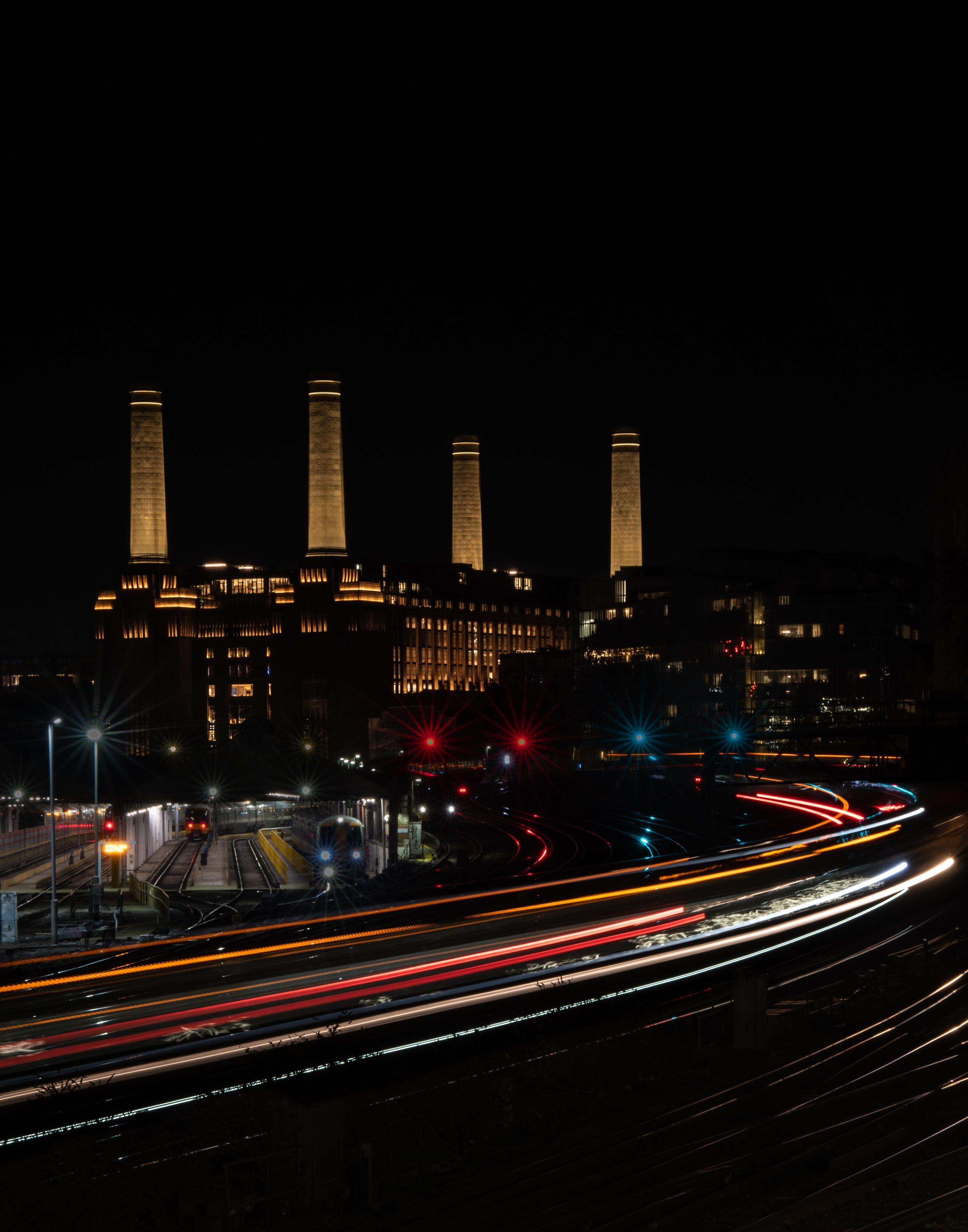 Night view of Battersea Power Station's illuminated chimneys with colorful light trails from trains curving across the foreground.