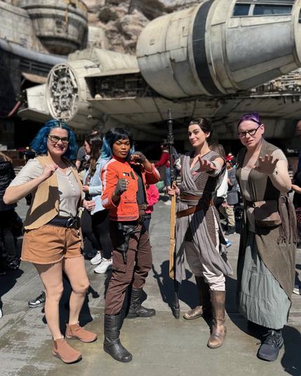 The image depicts four women standing in front of the iconic Millennium Falcon from Star Wars, posing for a photograph.

In the foreground, the woman on the left sports blue hair and is dressed in an orange top paired with brown shorts. The middle left woman also has blue hair and wears an orange jacket and brown pants. The middle right woman wears a gray dress complemented by brown boots, while the woman on the right has purple hair and is attired in a long brown coat over a white shirt and black pants.

The background of the image features the Millennium Falcon, which appears to be situated within a theme park or similar setting.