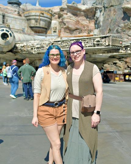 The image depicts two women standing together in front of a large spaceship, likely from the Star Wars franchise. The woman on the left has blue hair and is dressed in a white shirt, brown vest, and orange shorts. Her counterpart on the right wears a tan top with a long brown cardigan over it, paired with a gray skirt.

In the background, several individuals are visible, including one person wearing a green jacket and another sporting a backpack. The overall atmosphere suggests that this photograph was taken at Disneyland's Star Wars: Galaxy's Edge theme park.