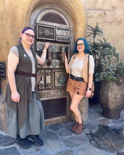 The image features two women posing in front of an ornate door, set against the backdrop of a stone wall.

The woman on the left is attired in a gray dress with a brown belt and black shoes. Her counterpart on the right sports blue hair and wears a white shirt paired with tan shorts and brown boots. Both individuals are positioned to the left of the door, which boasts an intricate design featuring various knobs and handles.

In the background, a stone wall provides a subtle yet elegant setting for the scene. The overall atmosphere suggests that the women are likely attending a themed event or celebration, given their attire and the ornate door behind them.